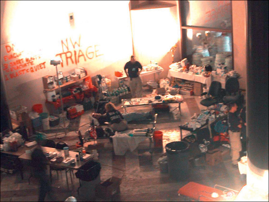 An interior view of an emergency triage station in New York in 2001, labeled N W Triage, with medical supplies, bottled water, I V fluids, equipment shelves, and personnel attending to patients.