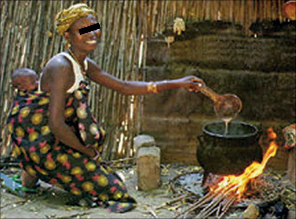 A person carries a baby on her back, tied with a cloth, while cooking outdoors over a stick fire using wooden utensils.