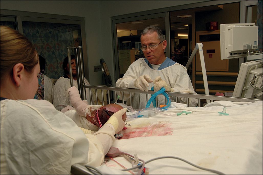 A doctor holds respiratory tubes while checking the patient. Another staff member tilts the patient’s head, the hand is bandaged, and two others observe at the bedside.