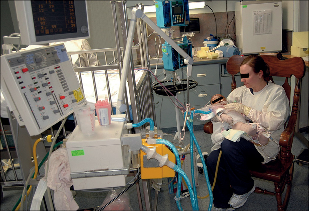 A healthcare professional holds a baby in her lap and presses the chest. They sit in a rocking chair surrounded by medical instruments, like in an operating room.