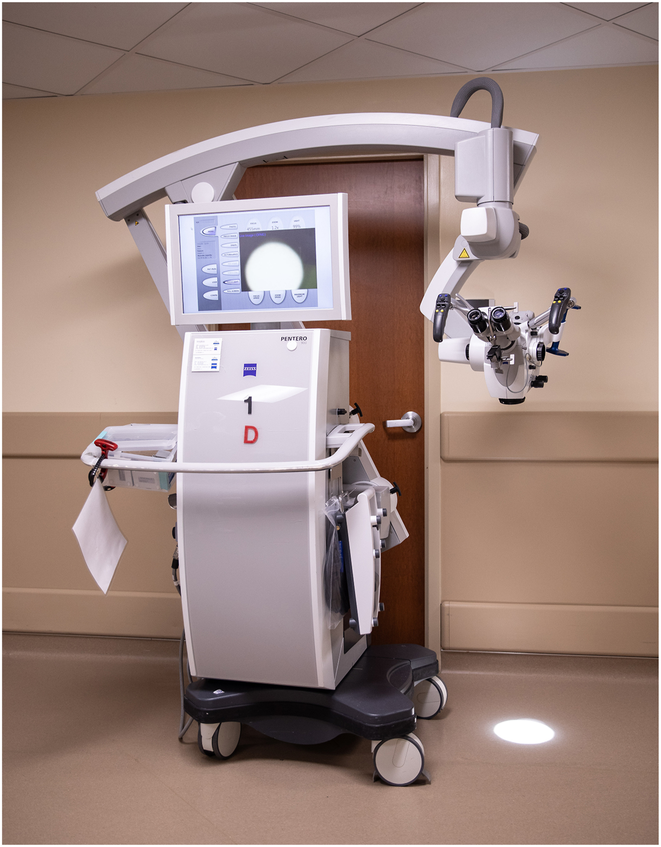 A large, wheeled surgical operating microscope with monitor, adjustable arm, and mounted optical equipment is positioned in a medical room.